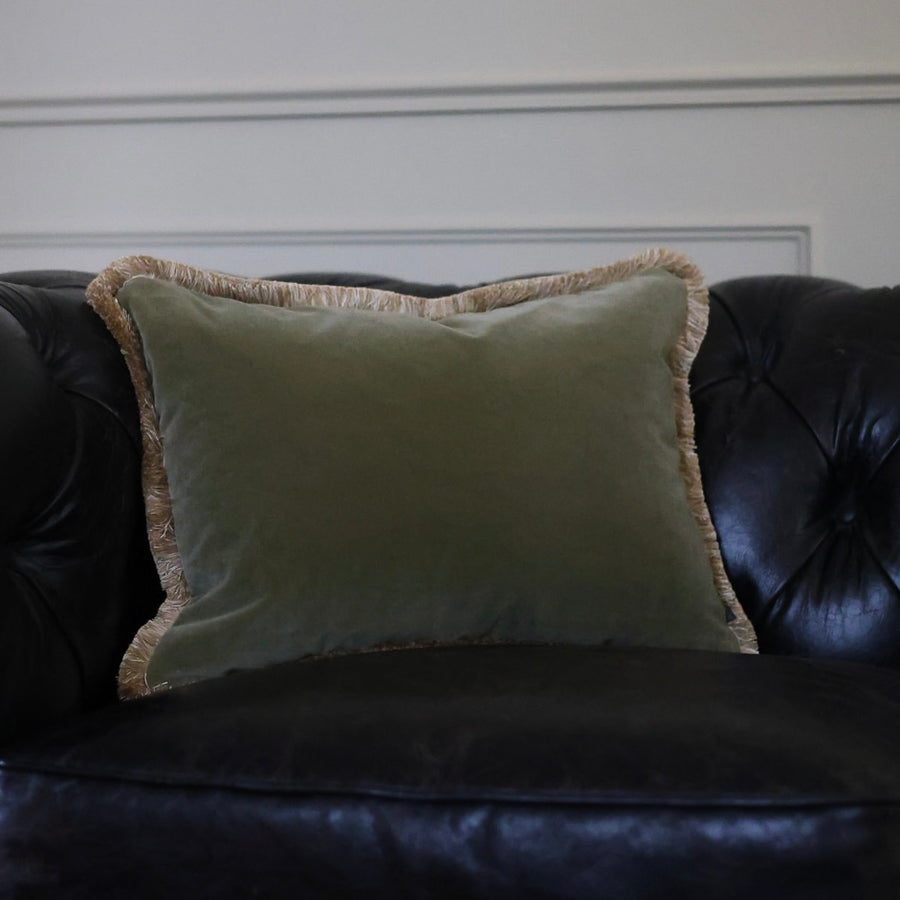 Rectangular moss green velvet cushion with gold fringe on a leather sofa.