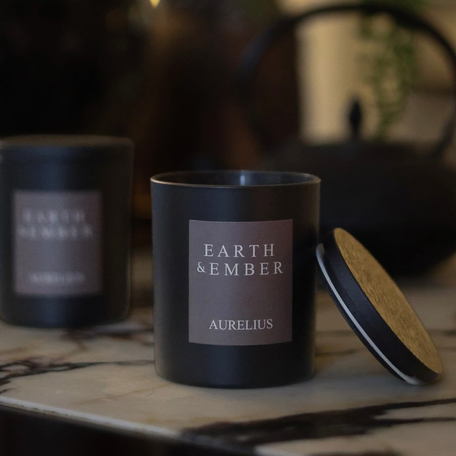 Earth & Ember Aurelius candle in a black vessel, with its wooden lid resting beside it on a marble surface.